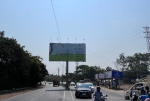 A large-format T-pole hoarding advertising a private events venue stands planted in the central median of NH-163, between Appa Junction and Chevella. The structure is clearly visible from the carriageway, constituting an illegal encroachment within the National Highway Right of Way.