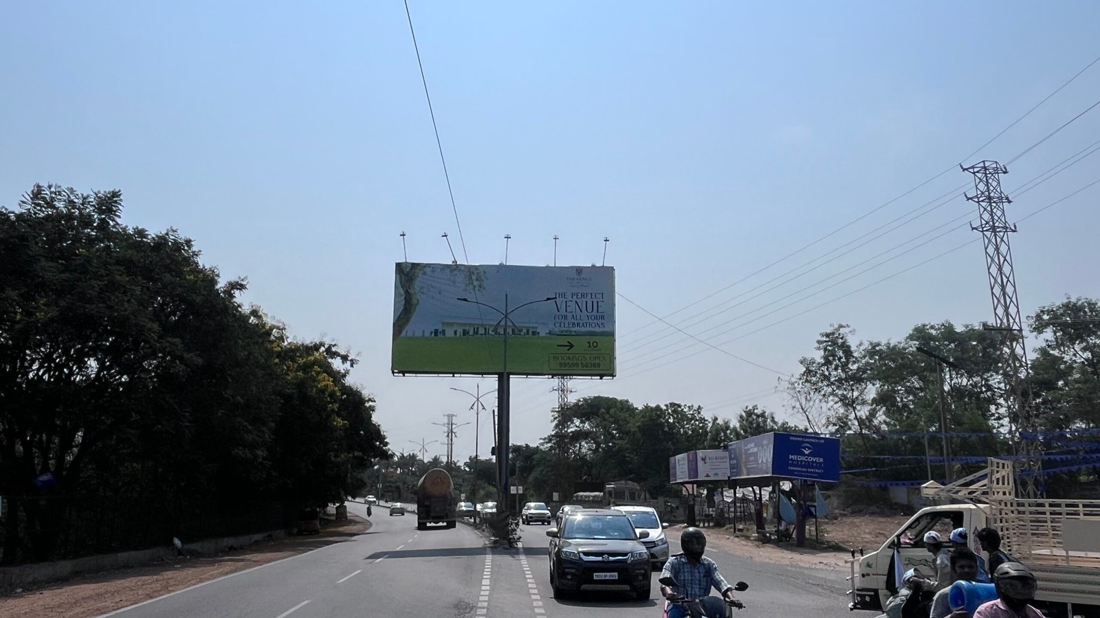 A large-format T-pole hoarding advertising a private events venue stands planted in the central median of NH-163, between Appa Junction and Chevella. The structure is clearly visible from the carriageway, constituting an illegal encroachment within the National Highway Right of Way.